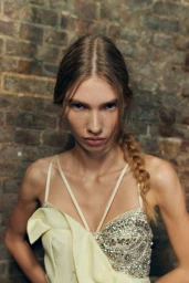 Close-up portrait of a model with braided hair wearing a pale yellow embellished dress against a brick wall background