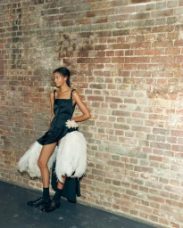Model leaning against a weathered brick wall wearing a black dress accented with white feathers
