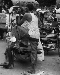 Black and white editorial photograph of men sitting and standing on a handcart in a busy street by Bharat Sikka for Lula Japan