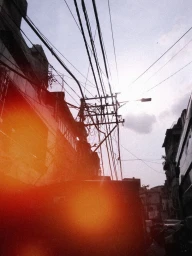 Urban street scene looking upward with silhouetted utility poles, tangled power lines, and buildings against a pale sky with light flare.