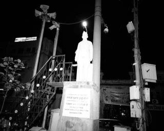 Monochrome night photograph of a statue on a plinth surrounded by utility poles, streetlights, and a metal staircase.