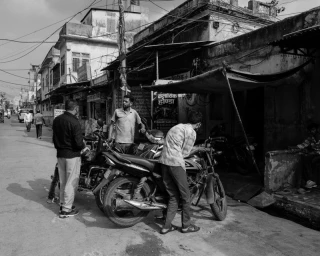 Black and white street scene featuring three men gathered around parked motorcycles on a narrow urban road.