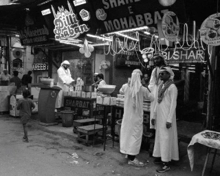 Monochrome street scene of men in traditional white thobes gathered around illuminated food and beverage stalls at night