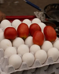 Close-up of fresh red tomatoes resting on top of a carton filled with white chicken eggs.