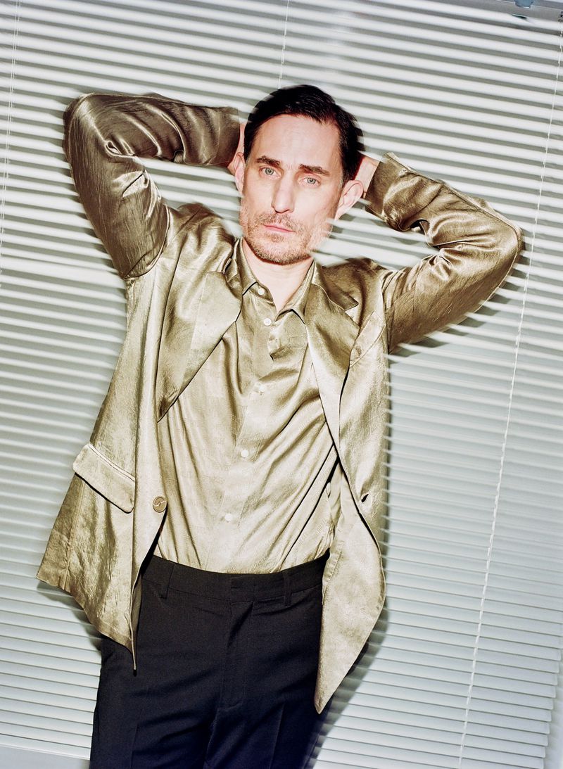Portrait of Clemens Schick in a shiny gold jacket and dark trousers standing against white window blinds