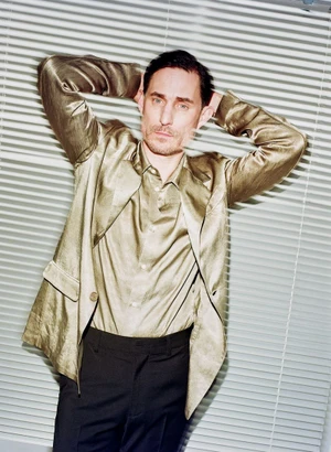 Portrait of Clemens Schick in a shiny gold jacket and dark trousers standing against white window blinds