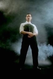Man standing in smoke wearing a white shirt and black trousers against a dark background for Sleek Magazine