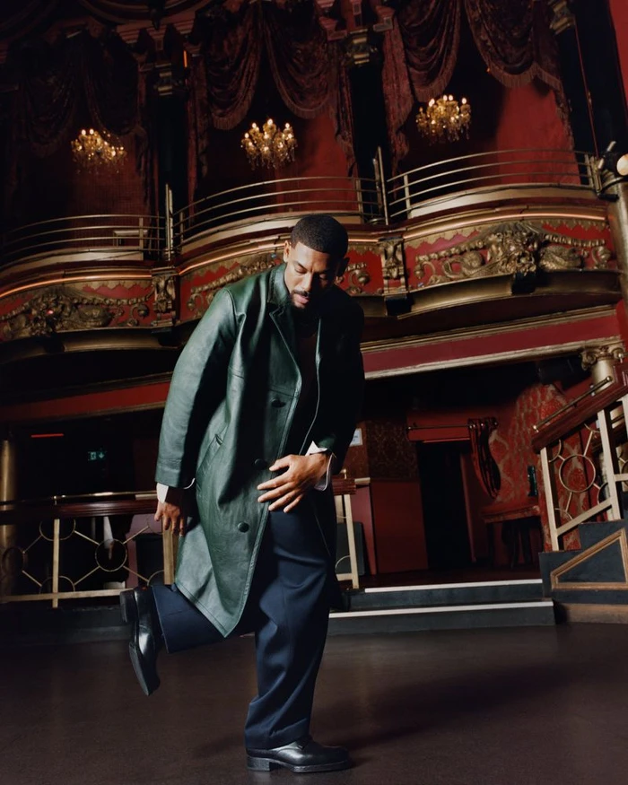 Aaron Pierre wearing a dark green leather coat and dark trousers inside an ornate theatre for Esquire UK