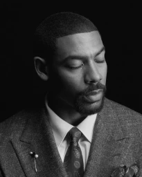 Portrait of actor Aaron Pierre with closed eyes wearing a tweed suit and patterned tie against a black background.