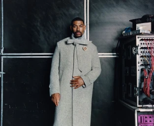 Actor Aaron Pierre wearing a textured grey coat with a bow collar standing before an industrial backdrop for Esquire UK.