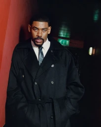 Portrait of actor Aaron Pierre wearing a black coat and tie in a dimly lit corridor for Esquire UK