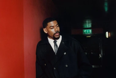 Aaron Pierre wearing a black coat and tie standing against a red wall for an editorial by Olivia Lifungula for Esquire UK