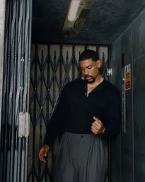 Actor Aaron Pierre standing in an elevator wearing a black henley shirt and grey trousers for Esquire UK.