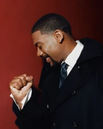 Portrait of actor Aaron Pierre smiling with a raised fist against a dark red background for Esquire UK photographed by Olivia Lifungula.