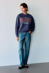 Model wearing a navy blue sweatshirt with Venice lettering and blue jeans against a grey studio wall