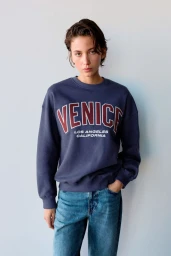 Model with short dark hair wearing a navy blue crewneck sweatshirt with 'Venice' lettering and blue jeans against a light background.