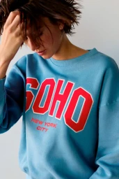 Model wearing a blue sweatshirt with red Soho New York City lettering against a plain wall