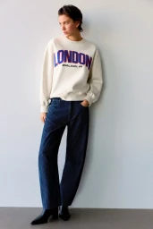 Model standing against a white wall wearing a white graphic sweatshirt and dark trousers, photographed by Louie Cobo for Pull&Bear.
