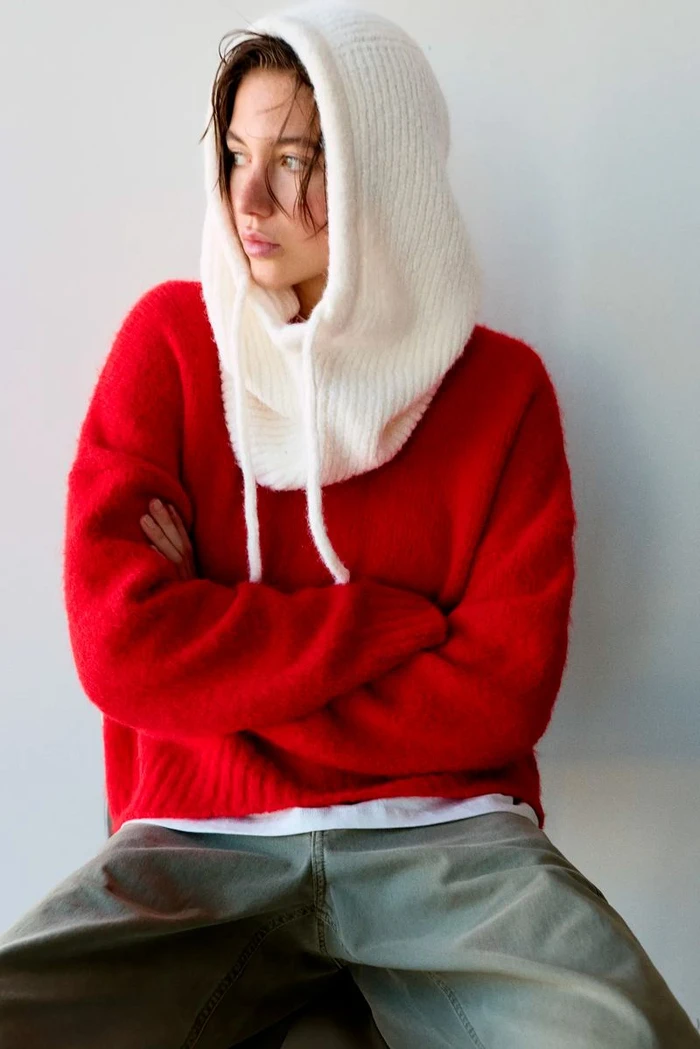 A woman wearing a white knit balaclava and a red sweater sitting against a plain white background for Pull&Bear.