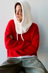 A woman wearing a white knit balaclava and a red sweater sitting against a plain white background for Pull&Bear.