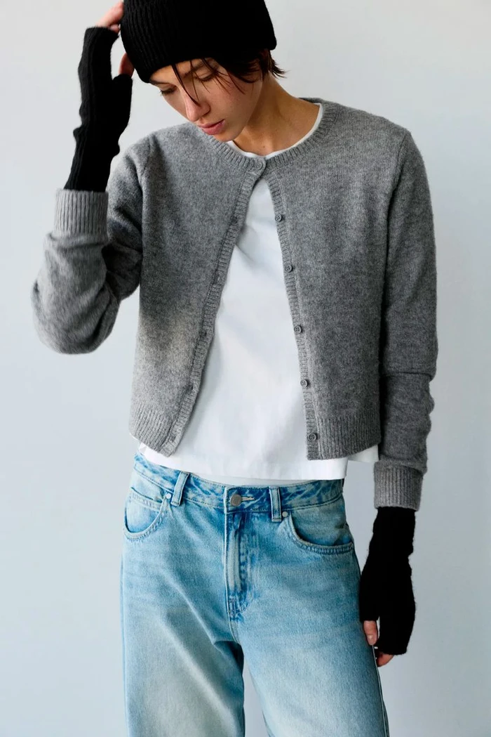 Model wearing a grey cardigan, white t-shirt, blue jeans, and black beanie against a white wall.