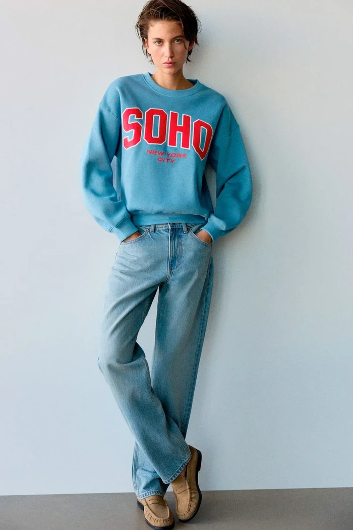 Model wearing a blue Soho sweatshirt and light wash jeans standing against a plain grey seamless background