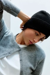 Model wearing a grey cardigan and black beanie adjusting hair against a white background