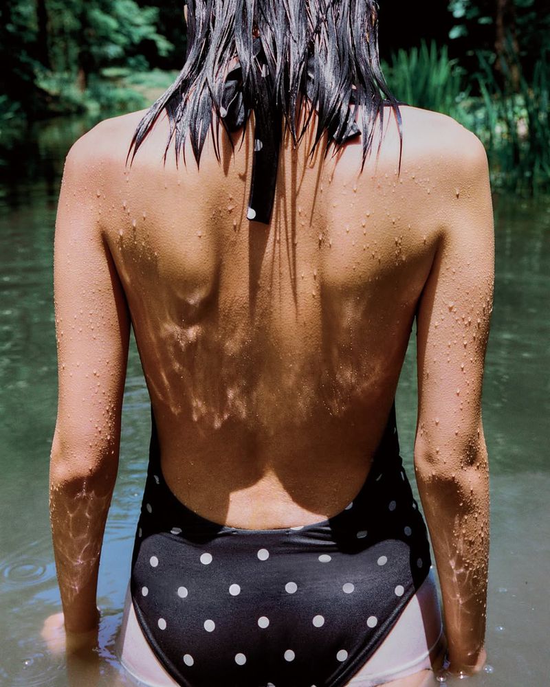 Back view of a woman in a polka dot swimsuit standing in water in a forest, sunlight dappling her skin, creating a serene outdoor scene.