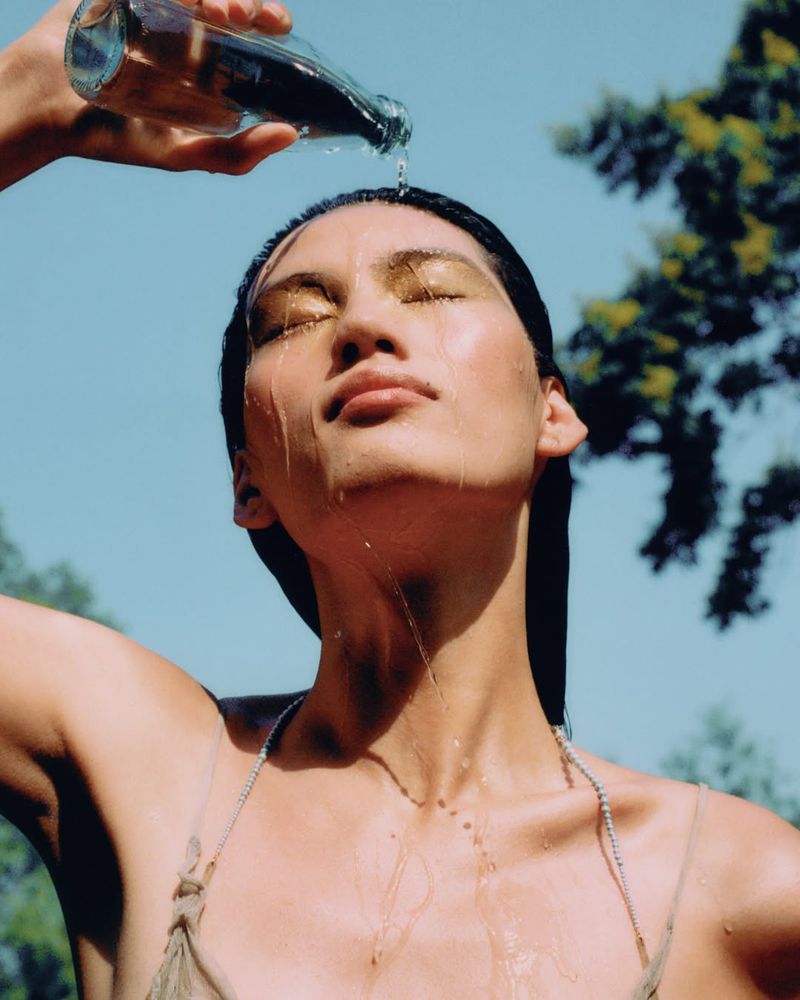 Woman outside with closed eyes is having water poured on her head from a glass bottle on a sunny day, with green trees visible.