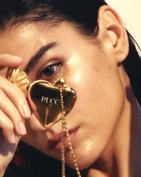 Close-up portrait of a woman holding a gold heart-shaped locket bearing the Pucci logo near her face.