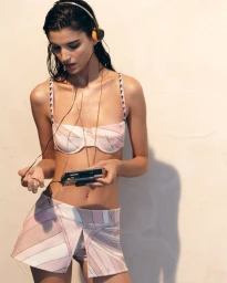 Model wearing patterned pink bikini and shorts listening to a portable cassette player with headphones against a neutral wall.