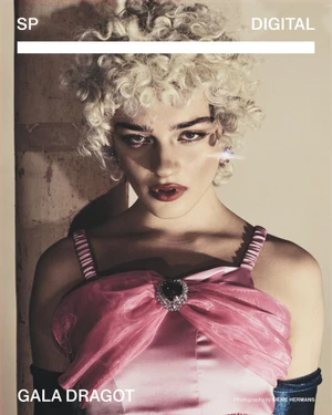 Model with blonde curly hair wearing a pink dress with a heart brooch and dark gloves against a pale wall.