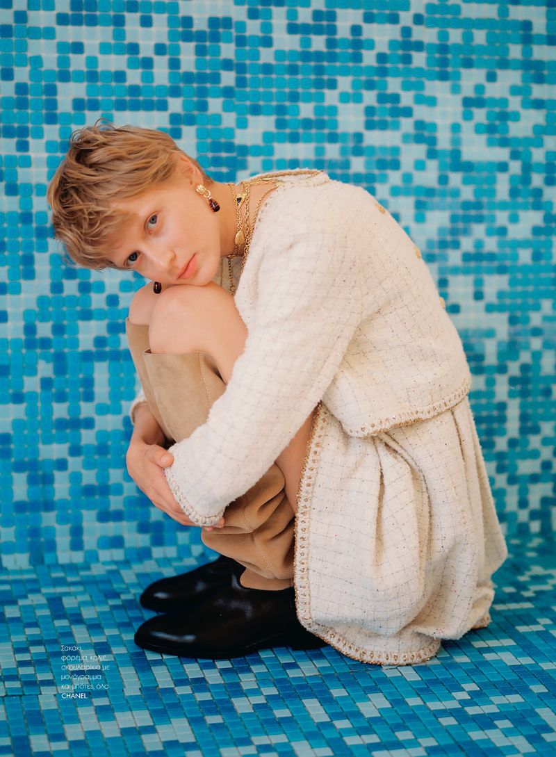 Fashion shot of a fair-skinned woman in a tweed jacket and skirt, hugging her knees, against a blue tiled pool background.