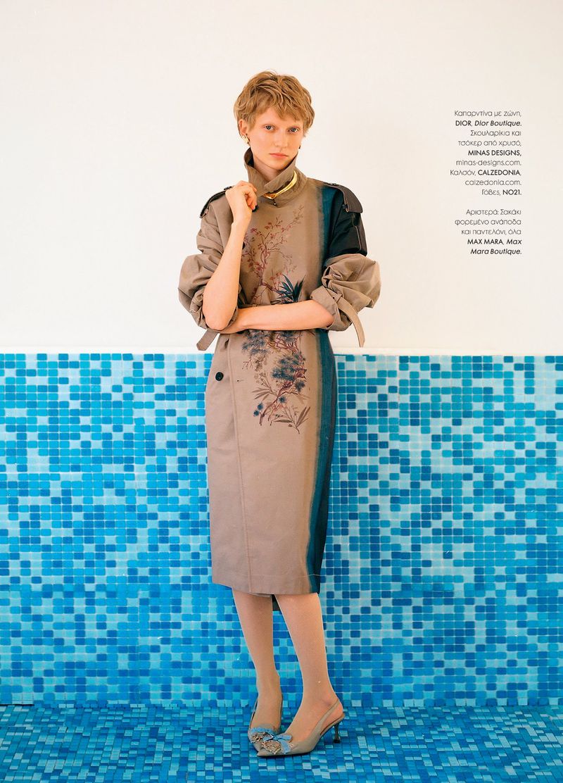 Fashion editorial shot of a woman in a beige and blue trench coat standing against a blue tile wall, with sheer hosiery and heels.