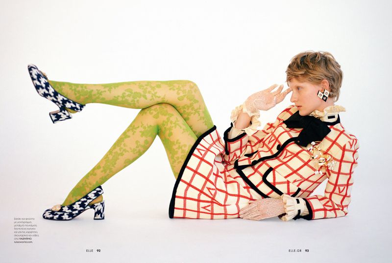 A blonde model poses in a patterned suit with green patterned tights and matching heels against a white backdrop in a studio shot.