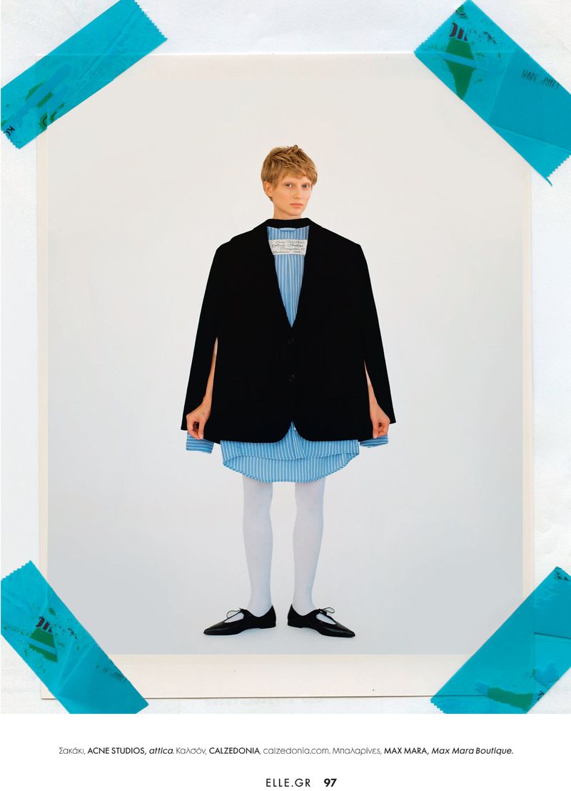 Fashion model in oversized black jacket and blue striped dress, white tights, black shoes, posing inside a white taped square.