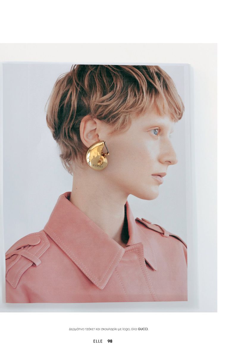 A side-profile view of a young adult female model with short blonde hair wearing a pink coat and large golden seashell earring indoors.