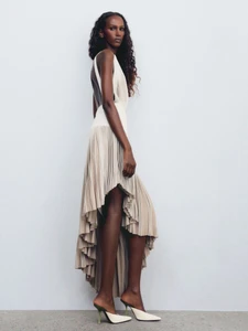 Model in a cream pleated high-low dress standing against a plain white studio background