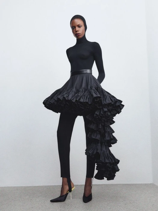 Model in all-black sculptural outfit with voluminous ruffled skirt and fitted top standing against plain wall