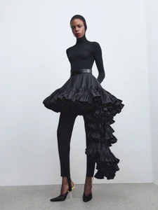 Model in all-black sculptural outfit with voluminous ruffled skirt and fitted top standing against plain wall