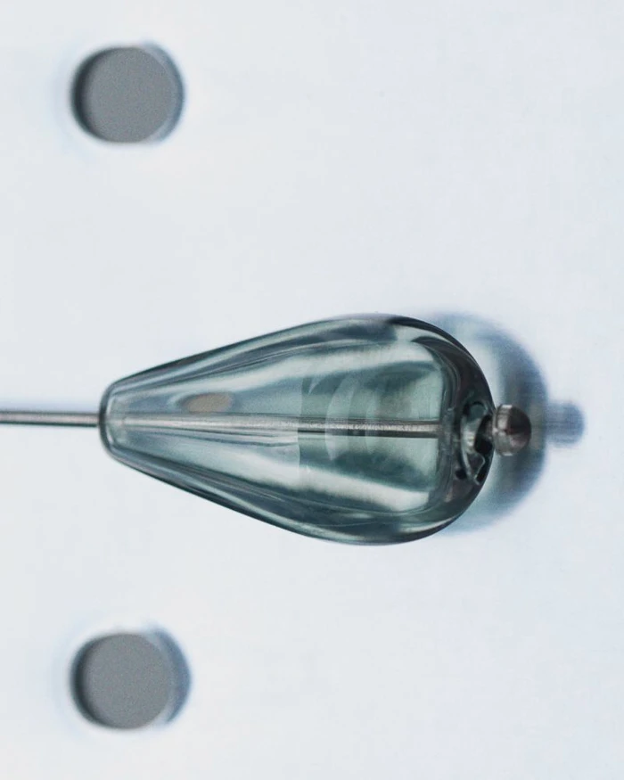 Close-up of a teardrop-shaped glass object pierced by a metallic pin against a light background.