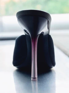Close-up rear view of a black high-heeled shoe featuring a stiletto heel with a gradient purple fade, set on a reflective surface.