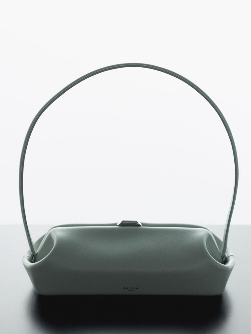 Minimalist studio shot of a sage green leather handbag with a high arched handle against a white and dark surface.