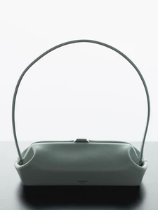 Minimalist studio shot of a sage green leather handbag with a high arched handle against a white and dark surface.