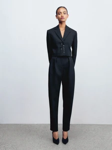 Model standing against a white backdrop wearing a black tailored blazer with decorative buttons and matching trousers.