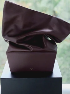 Dark burgundy leather handbag with slouchy sculptural form resting on a black rectangular pedestal against a soft green window view