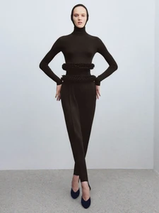 Model in a black turtleneck outfit with a hooded headpiece standing against a seamless grey backdrop.