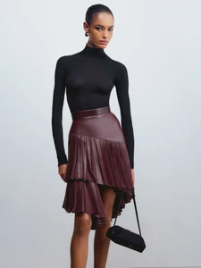Model in a black turtleneck and burgundy pleated leather skirt holding a black handbag against a seamless white background.