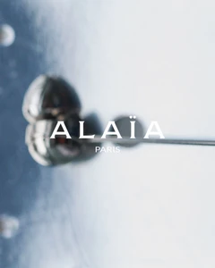Close-up abstract fashion shot with the Alaïa Paris logo centered over a blurred reflective metallic surface