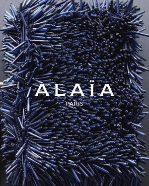 Close-up of a dark navy blue beaded handbag covered in dense fringe and spikes with the word ALAIA PARIS printed in white across the center